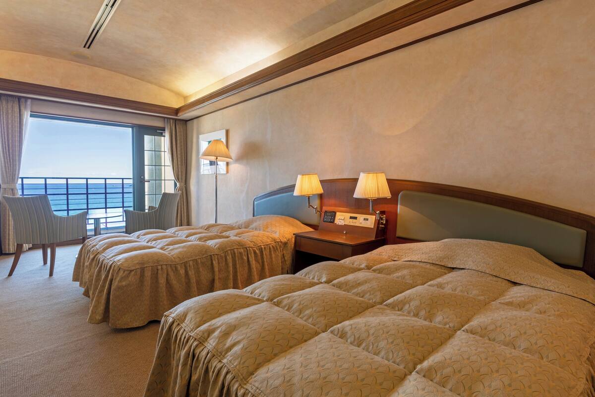 A hotel room with two beds, a desk, a chair, and a view of the sea.