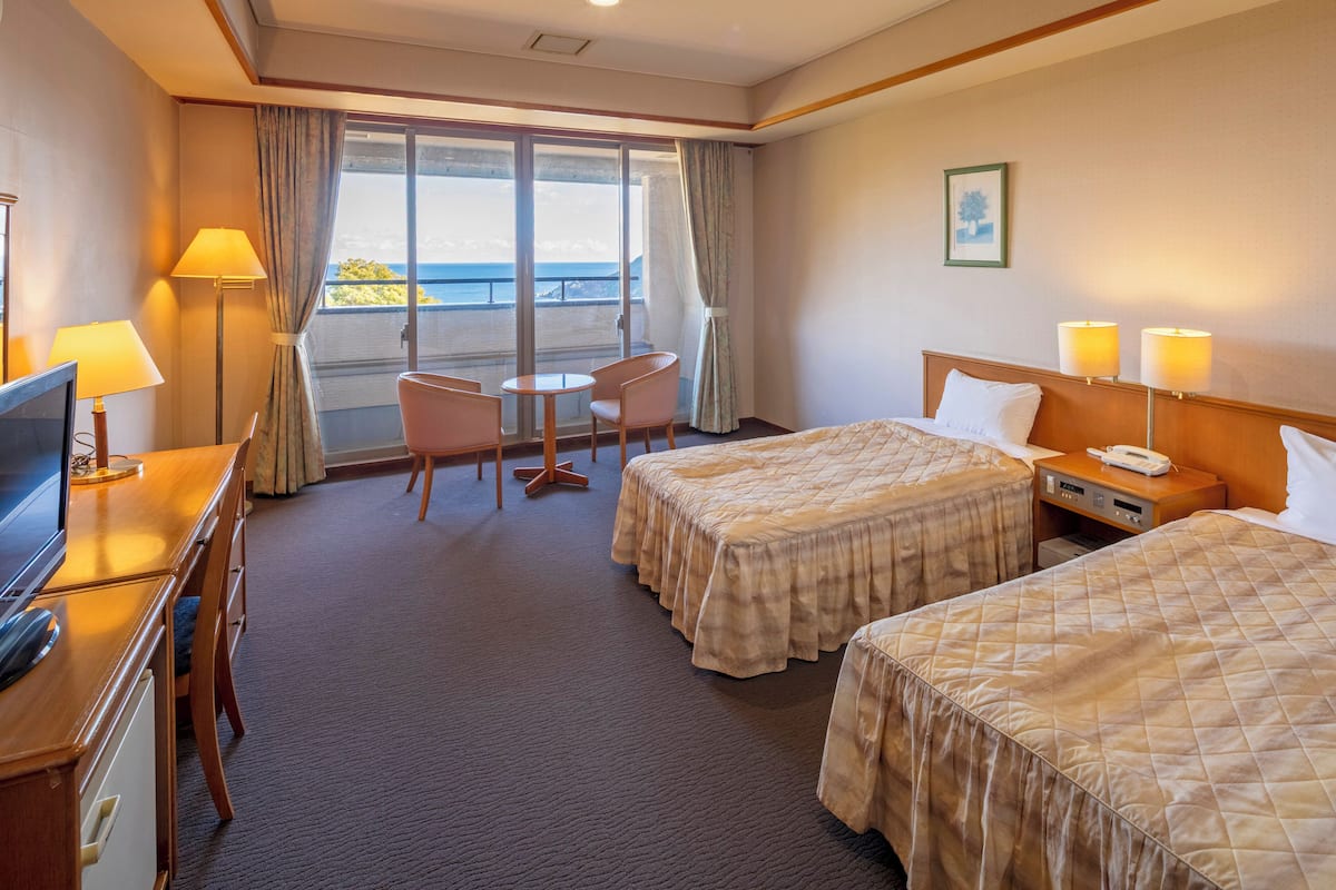 A hotel room with two beds, a desk with a TV, a chair, a small table, and a balcony with curtains.