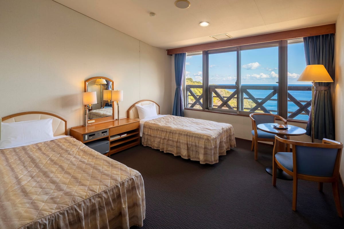 A hotel room with two beds, a small table with chairs, a desk with a lamp, and a large window with a view of the sea.