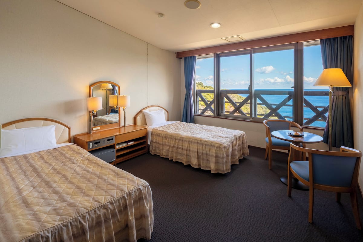 A hotel room with two beds, a desk, a chair, a table, and a large window with a view.