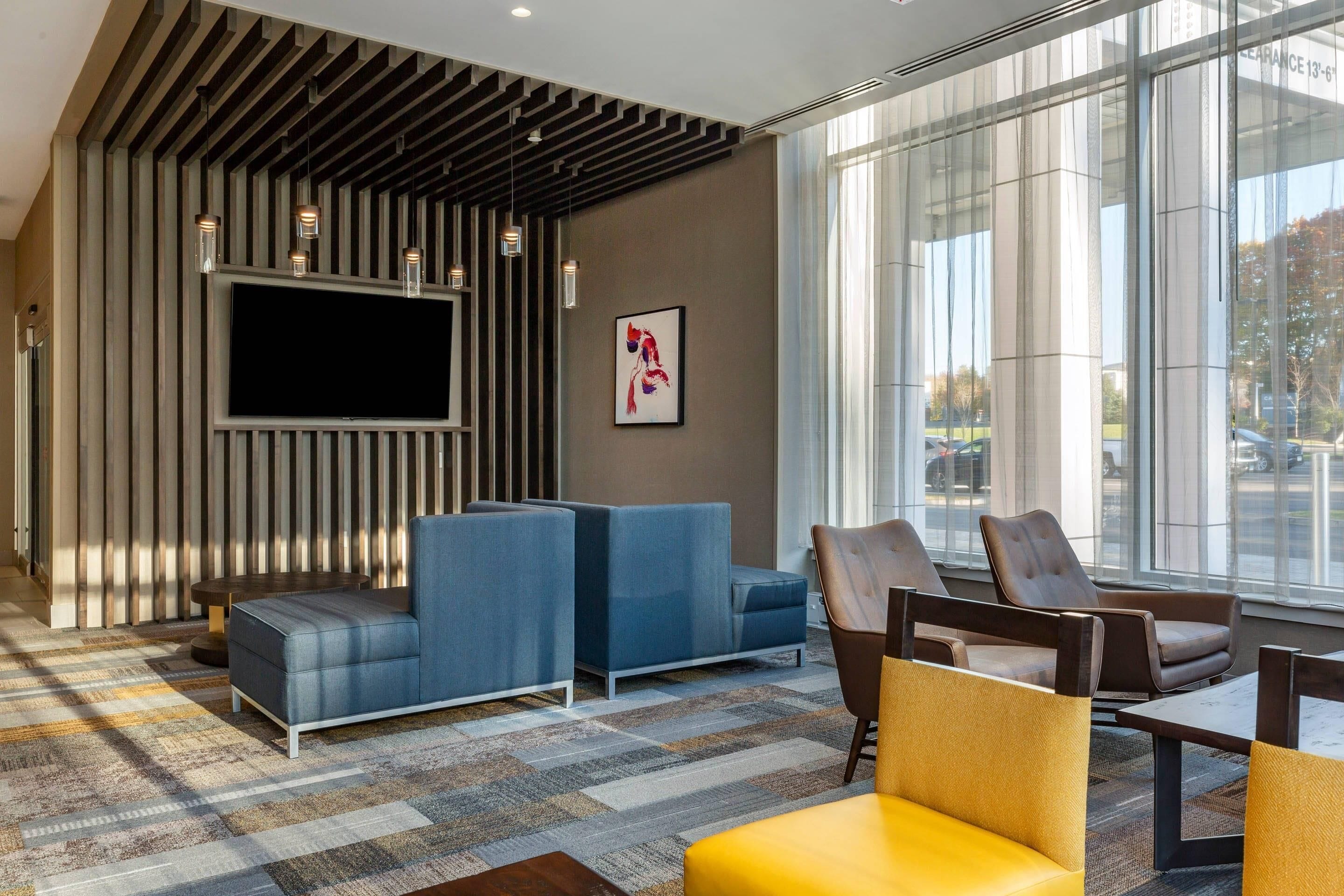 A modern lobby with a large flat-screen TV, seating areas with blue and brown chairs, and a patterned carpet.