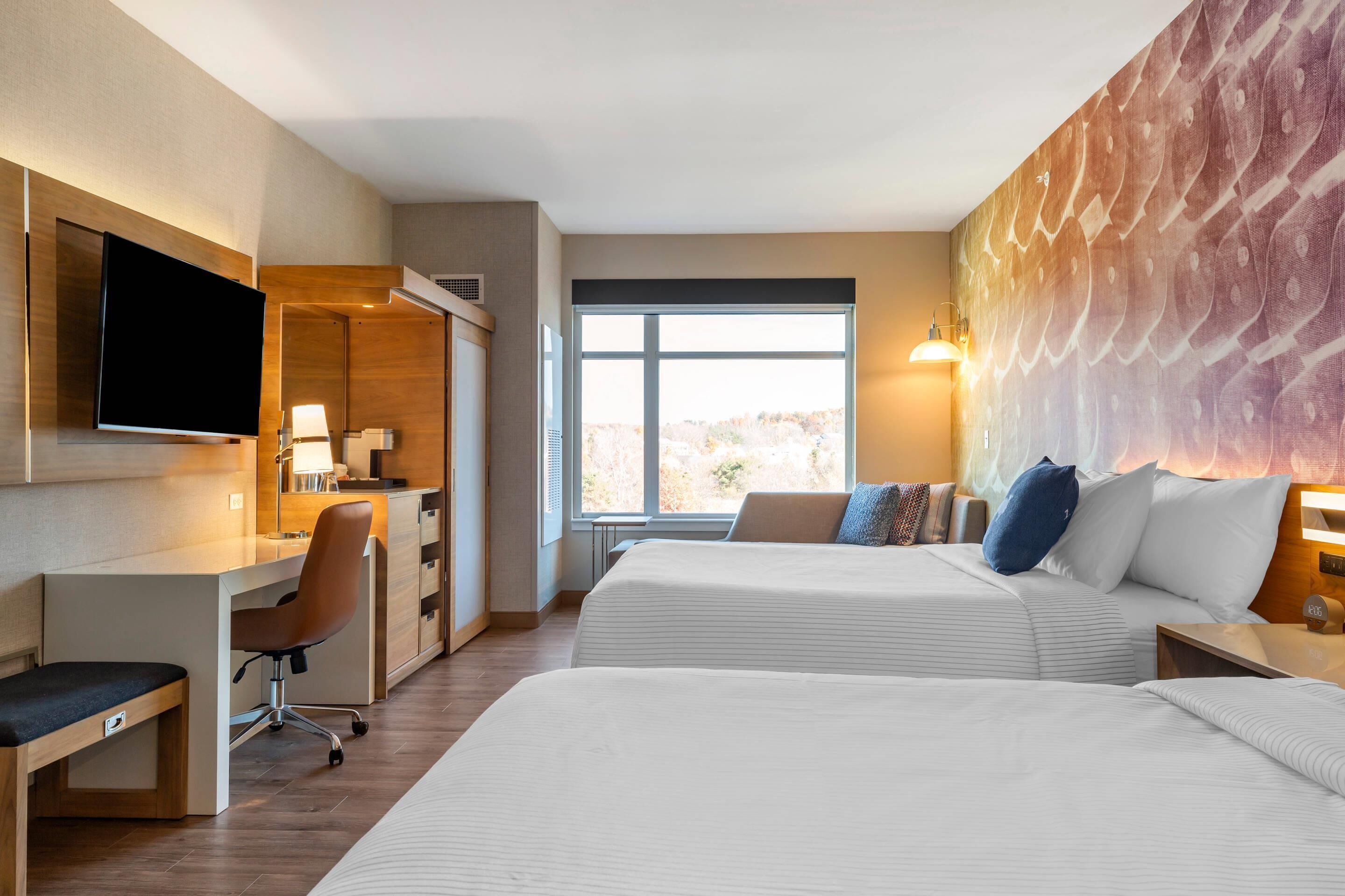 A hotel room with two beds, a desk, a TV, and a large window.