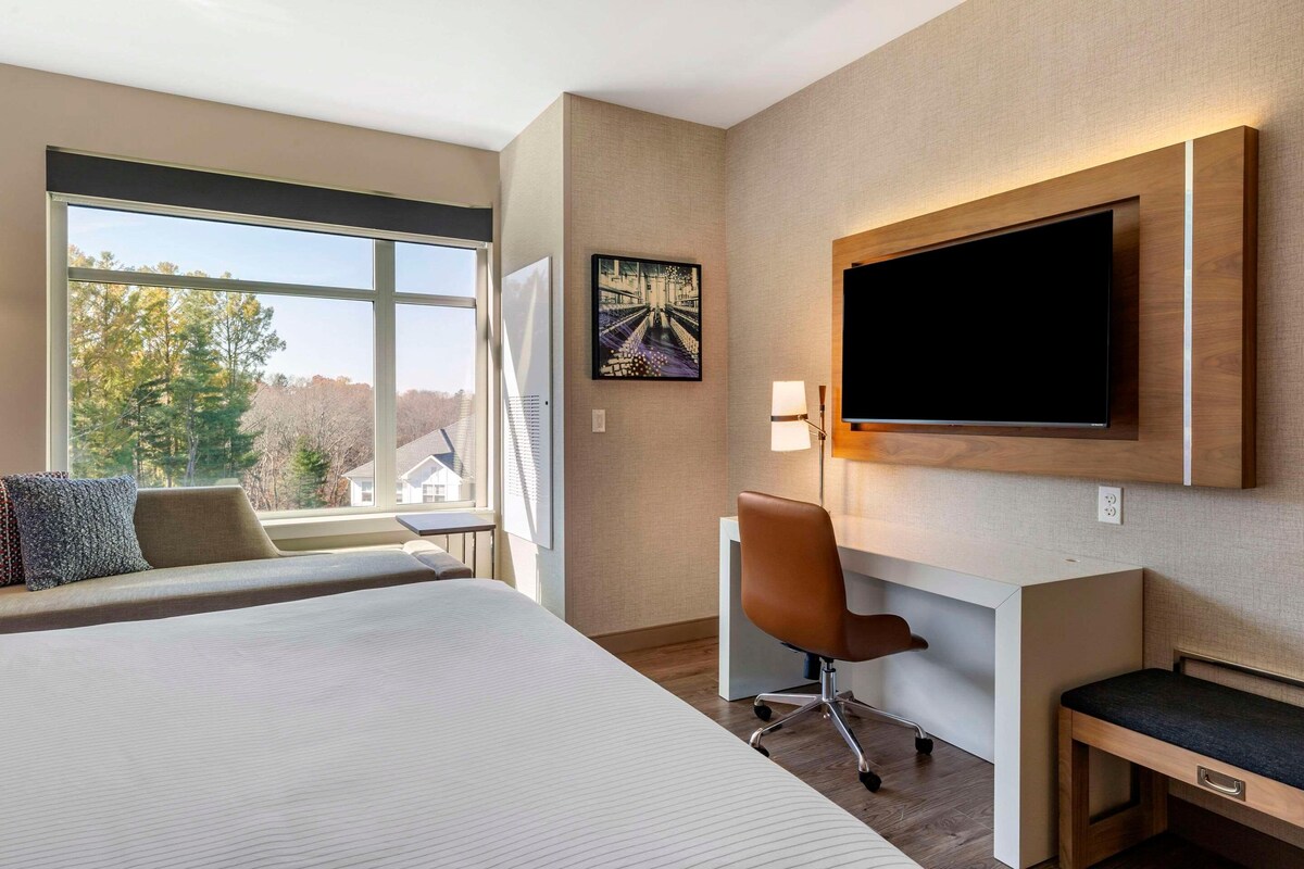 Habitación de hotel con una cama grande, un escritorio con silla, televisión y ventana con vistas a árboles y edificios.