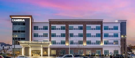 A multi-story hotel with a prominent CAMBRIA sign, a parking lot with several cars, and a clear sky.