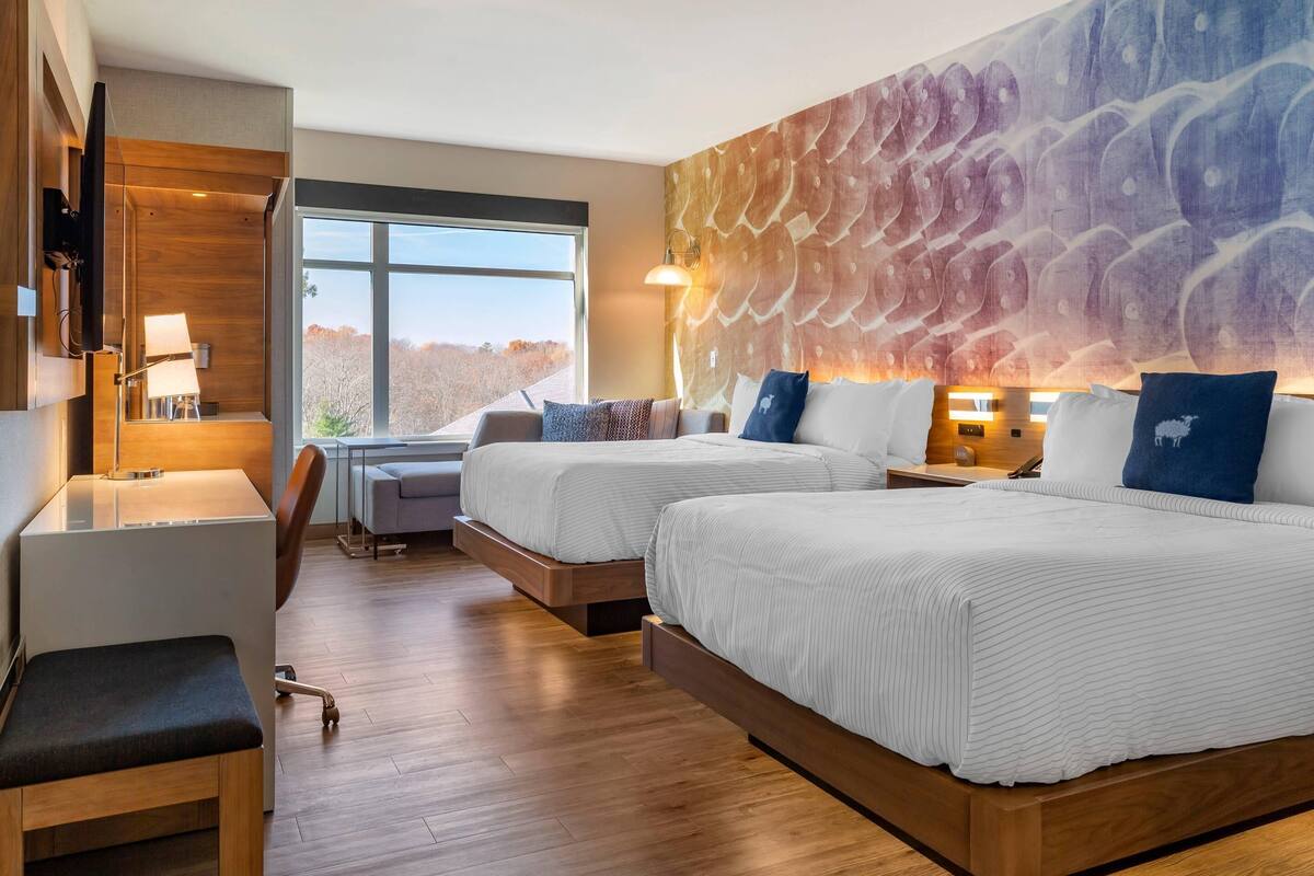A hotel room with two beds, a desk, a chair, a large window, and a wall mural.