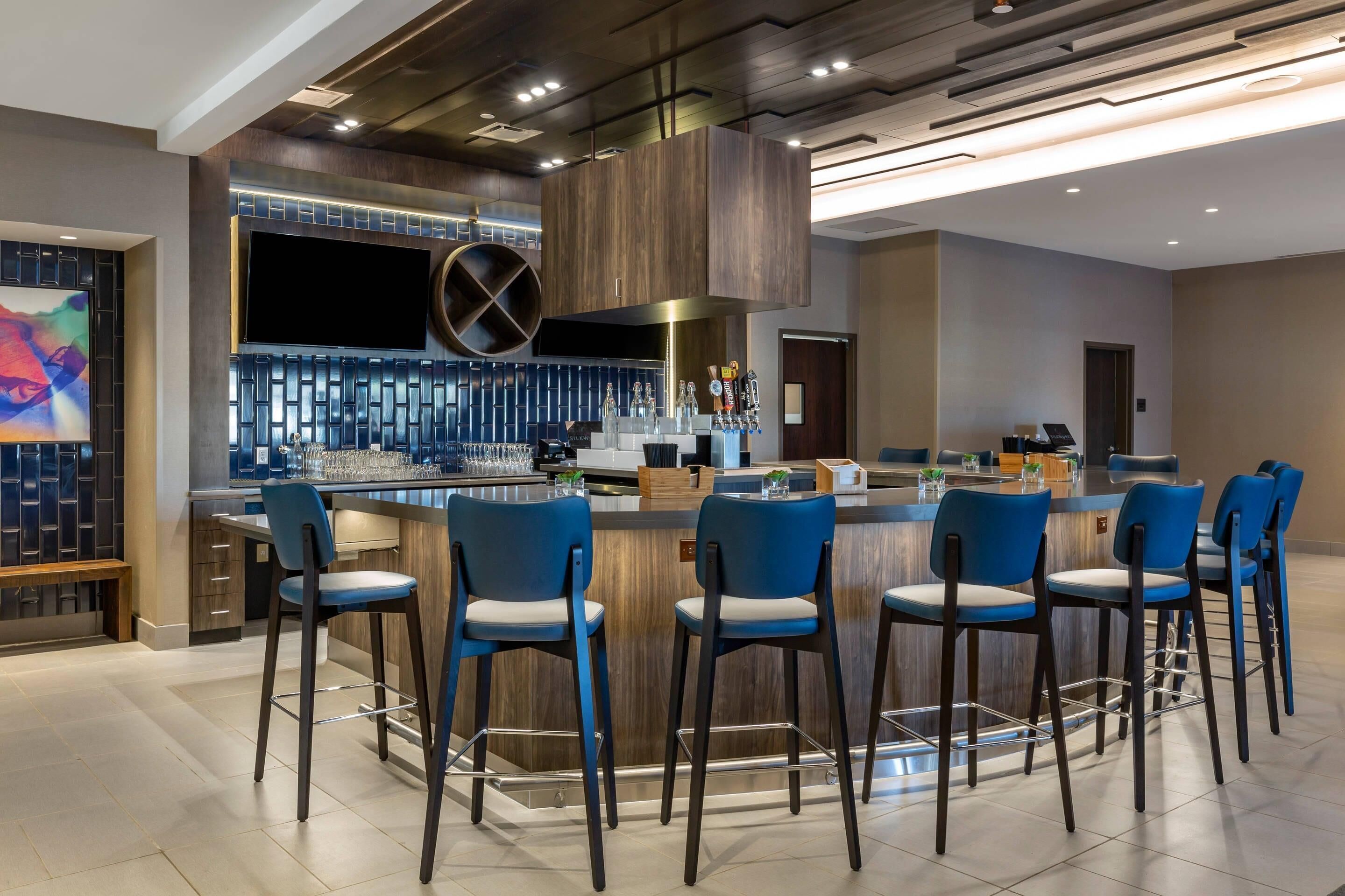 Zona de bar moderna con barra central, varios taburetes y una pantalla grande sobre la barra.