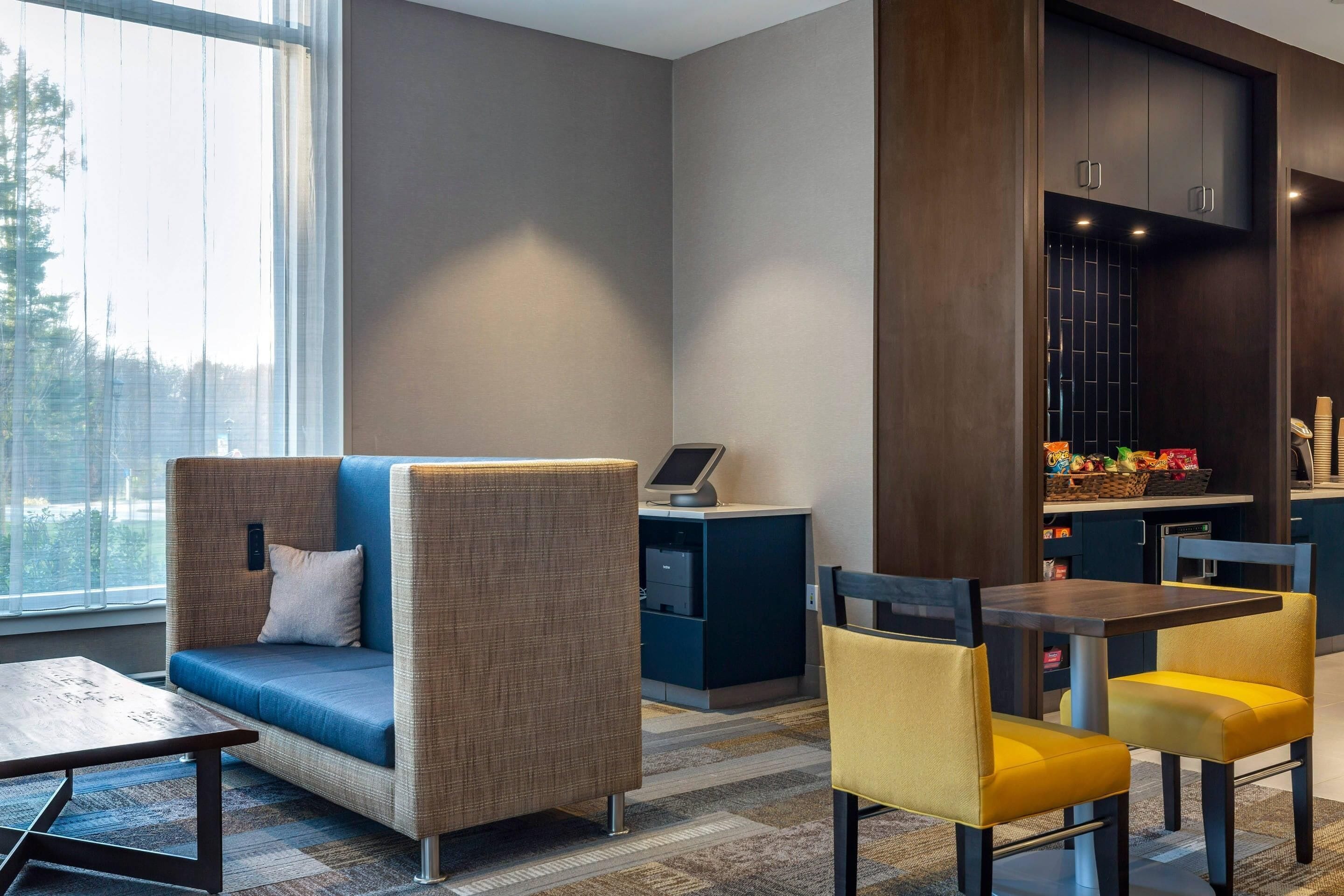 A modern hotel lobby with a sofa, chairs, a desk with a computer, and a view of the outdoors.