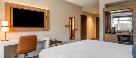 A hotel room with a large bed, a desk with a chair, a television mounted on the wall, a microwave, and a small kitchenette.