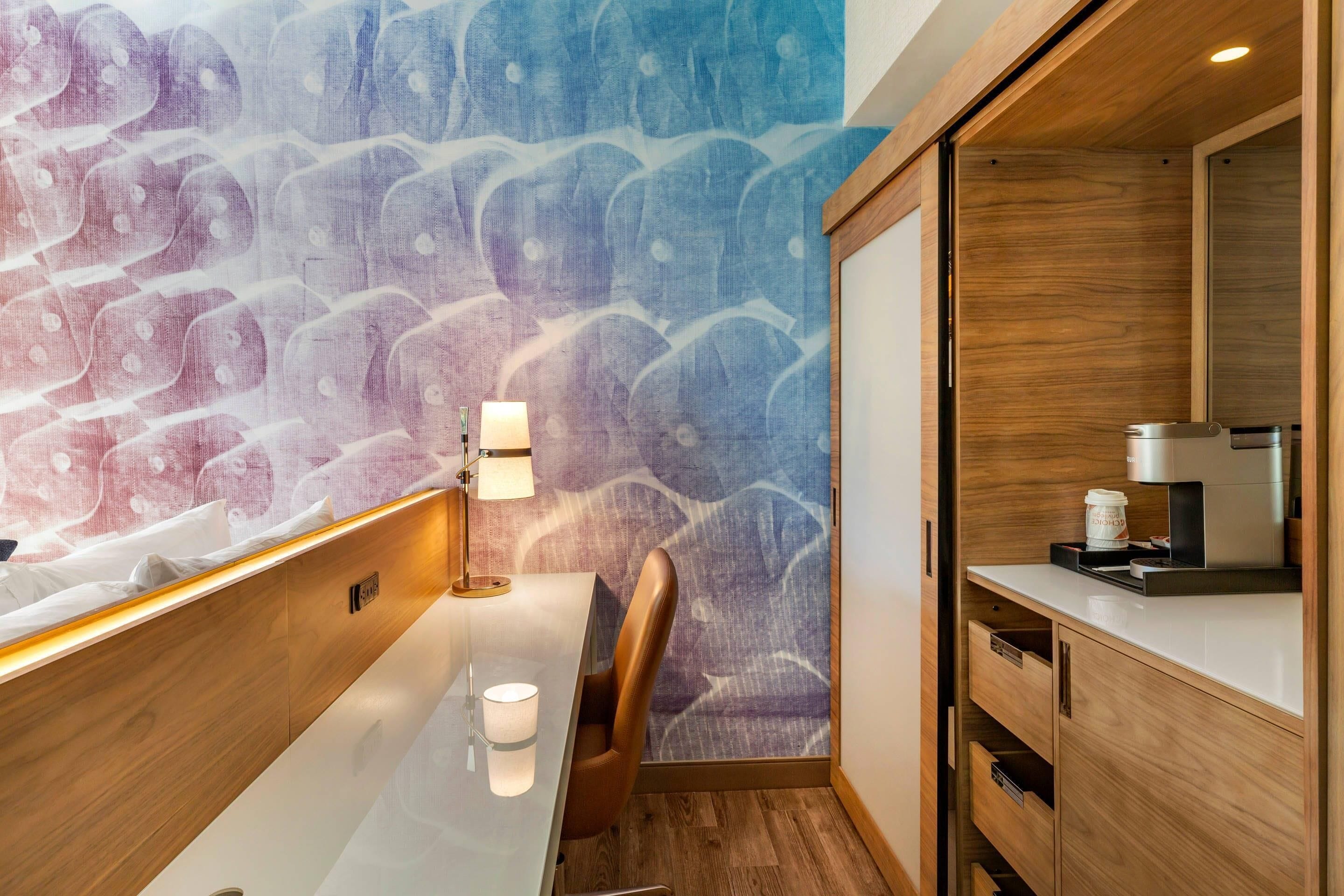 A modern hotel room with a wooden desk, a chair, a lamp, a coffee machine, and a patterned wall.