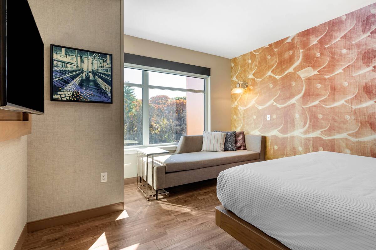 A modern hotel room with a large bed, a sofa, a TV mounted on the wall, a window with a view of trees, and a textured wall.