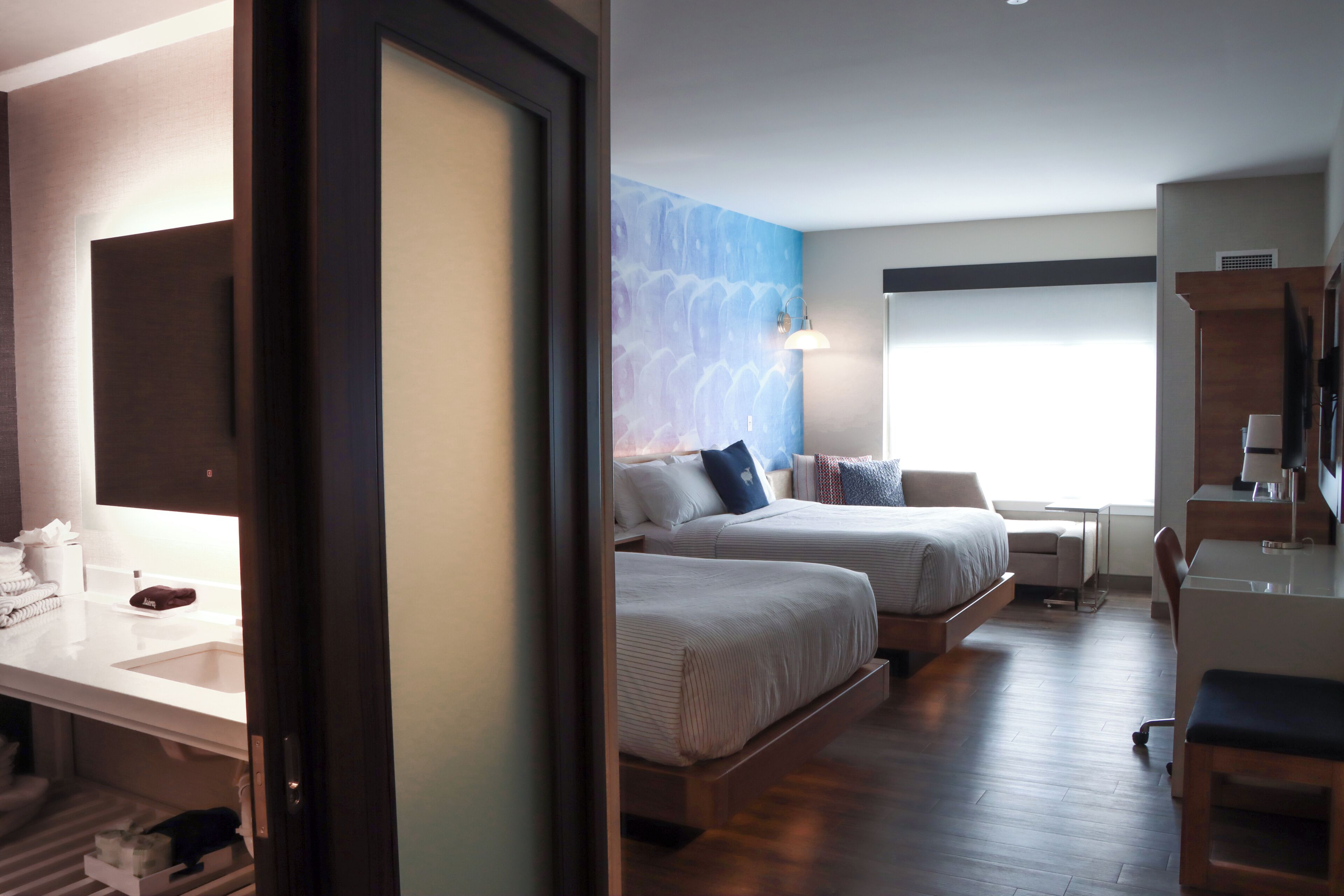A hotel room with two beds, a desk, a chair, a desk lamp, and a large window.