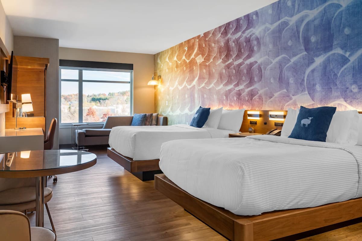 A hotel room with two beds, a desk, a chair, a lamp, and a large window.