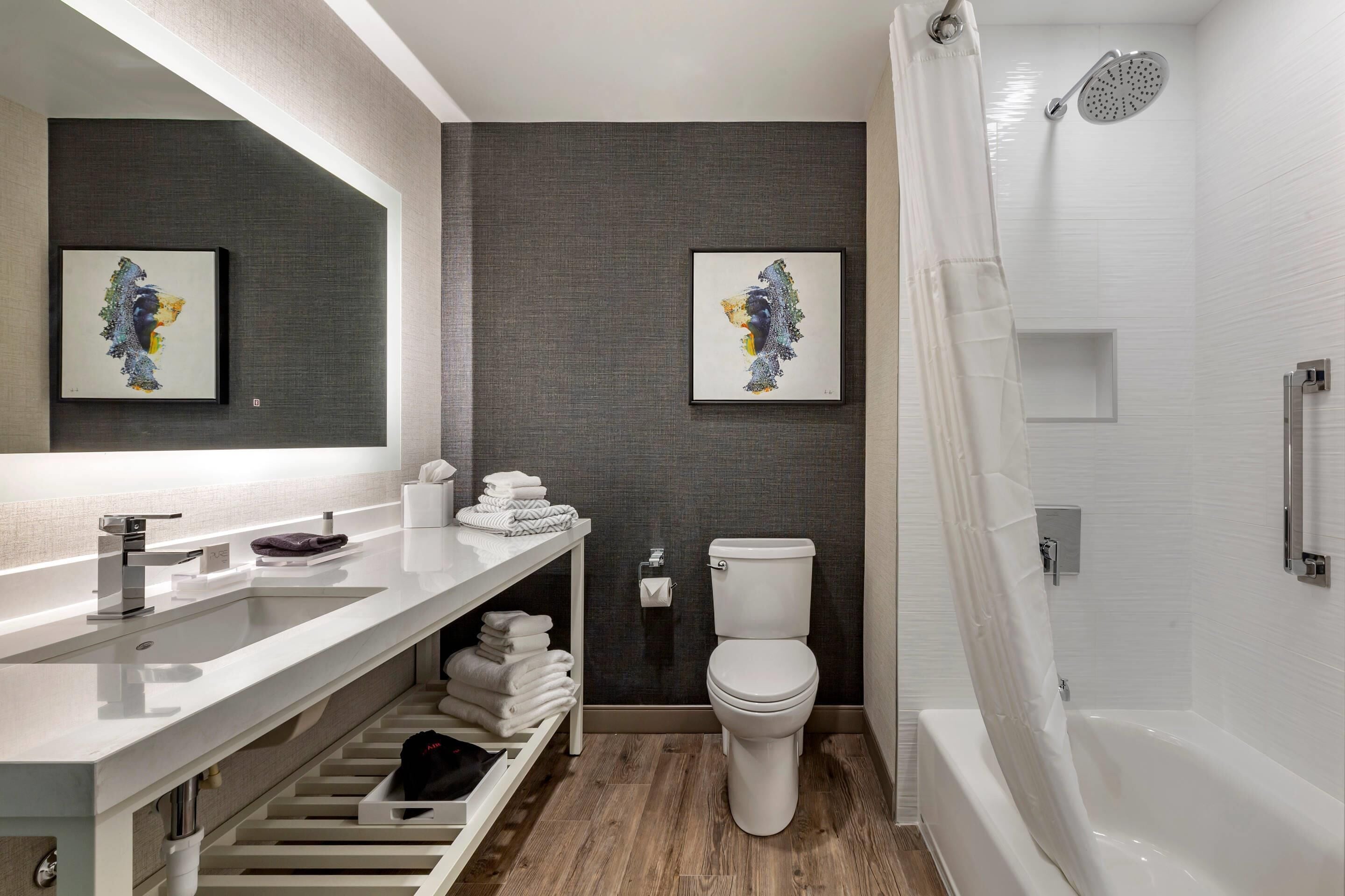 Ein modernes Badezimmer mit einem weißen Waschbecken, einer Toilette und einer Badewanne. An den Wänden hängen zwei gerahmte Kunstwerke, ein Handtuchhalter und ein Regal mit gefalteten Handtüchern.