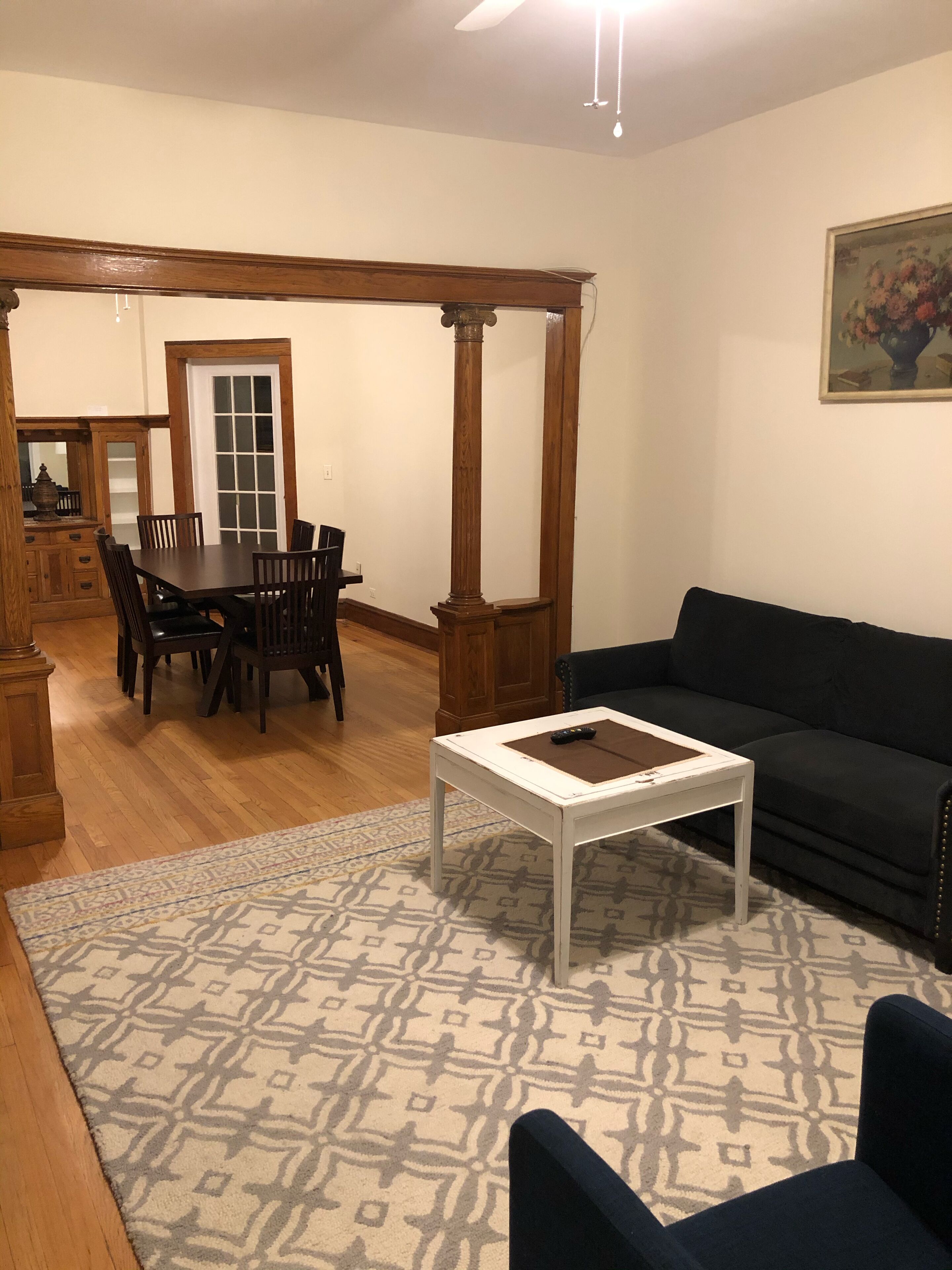 A living room with a dark-colored sofa, a white coffee table, a patterned rug, wooden flooring, and a dining area with a wooden table and chairs.