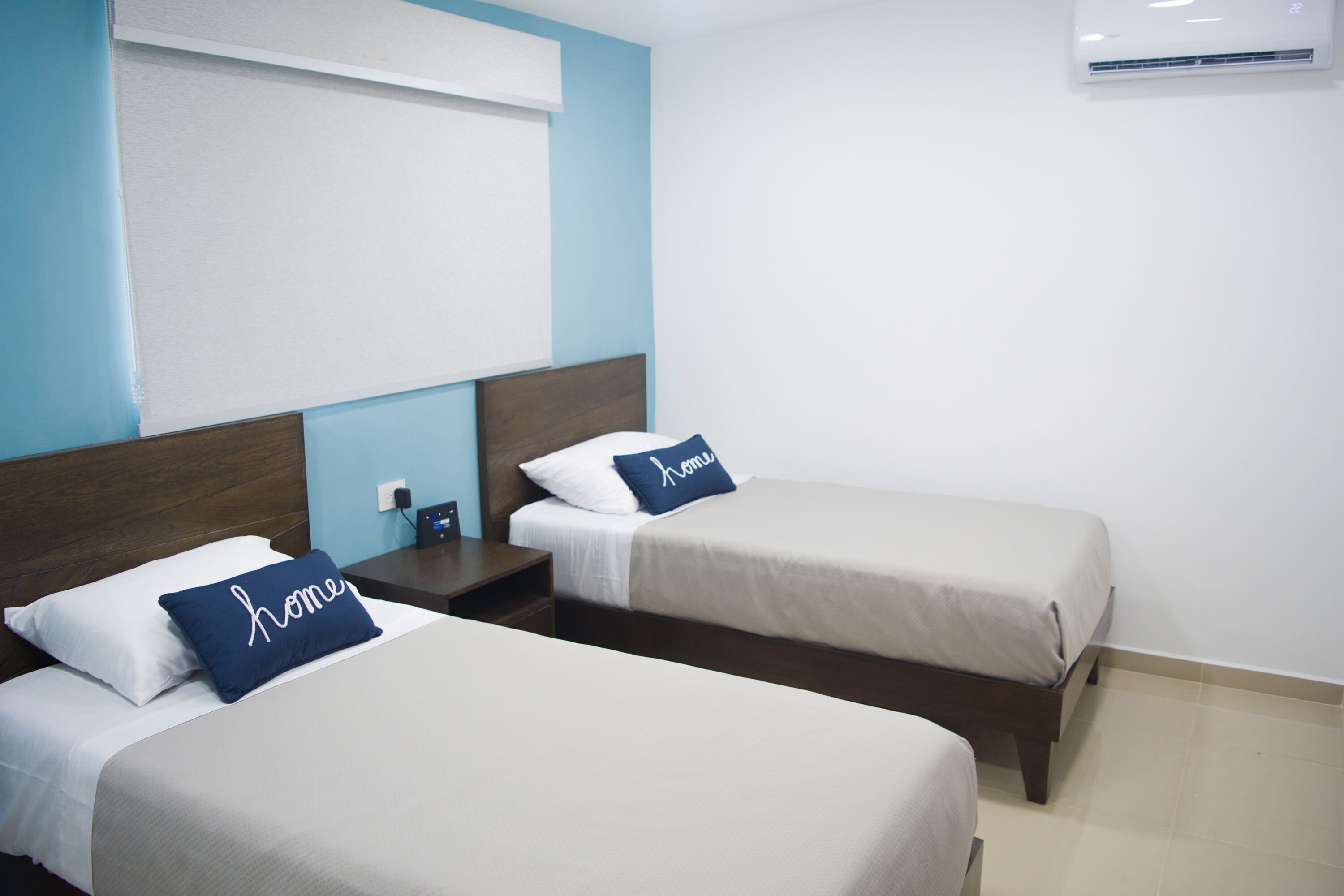 Quarto de hotel com duas camas de solteiro, mesa de cabeceira, ar-condicionado fixado na parede e uma janela com persianas.