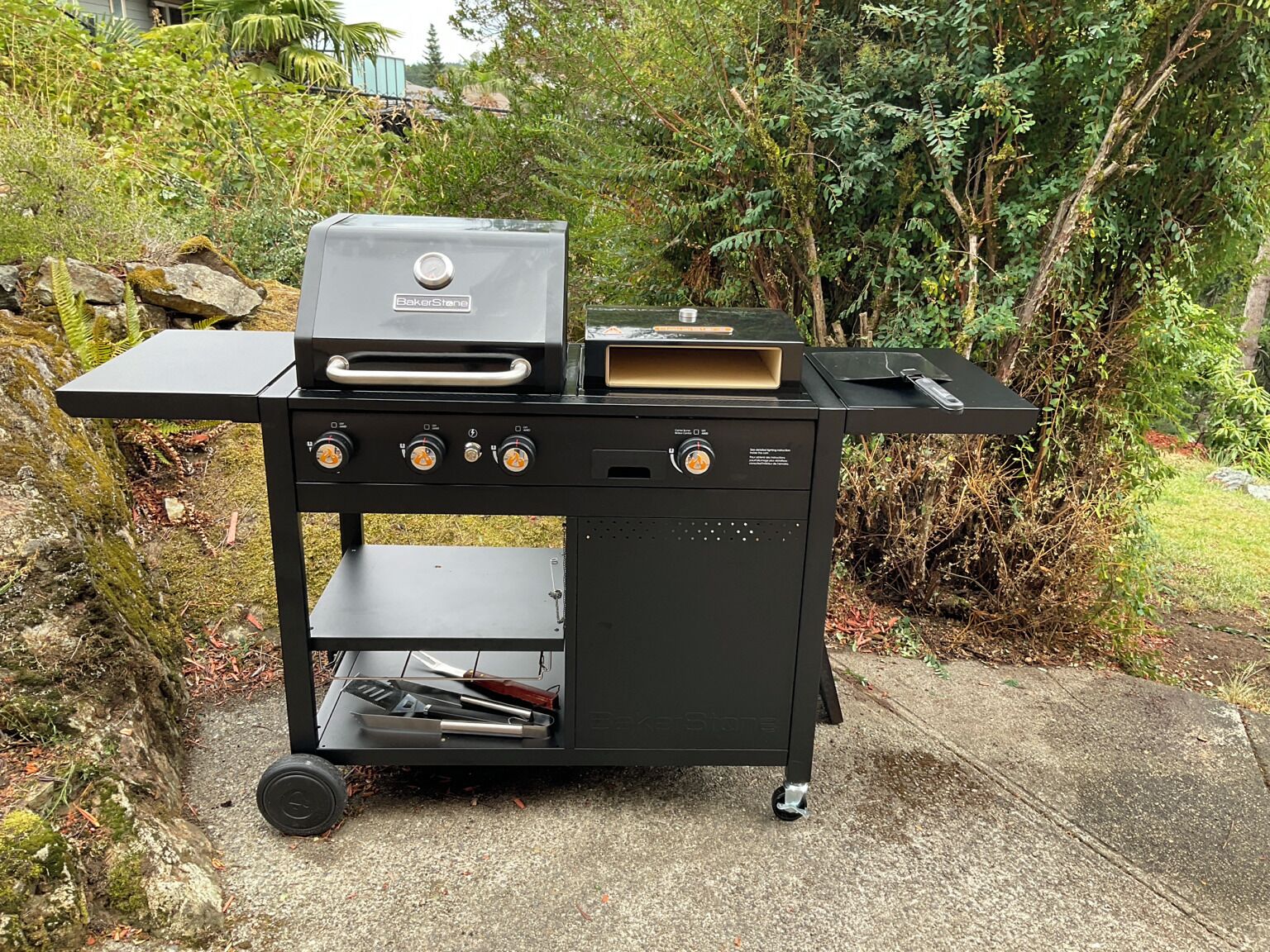 Een draagbare zwarte grillwagen met ingebouwde oven en een vlakke grillplaat, voorzien van meerdere bedieningsknoppen en een opbergvak.