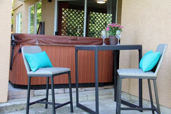 A hot tub with a wooden cover, two chairs with turquoise cushions, and a table with two wine glasses and a potted plant.