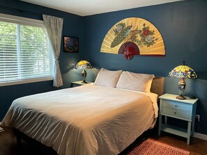 A bedroom with a bed, two bedside tables with lamps, a window with curtains, and a wall-mounted decorative piece.