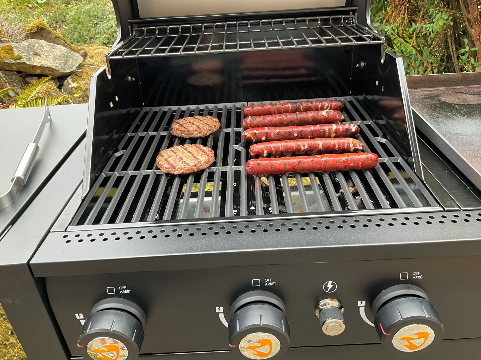 A grill with four sausages and two burgers cooking on the grates.