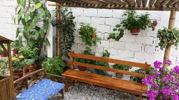 A wooden bench under a bamboo pergola, surrounded by potted plants and a white brick wall.