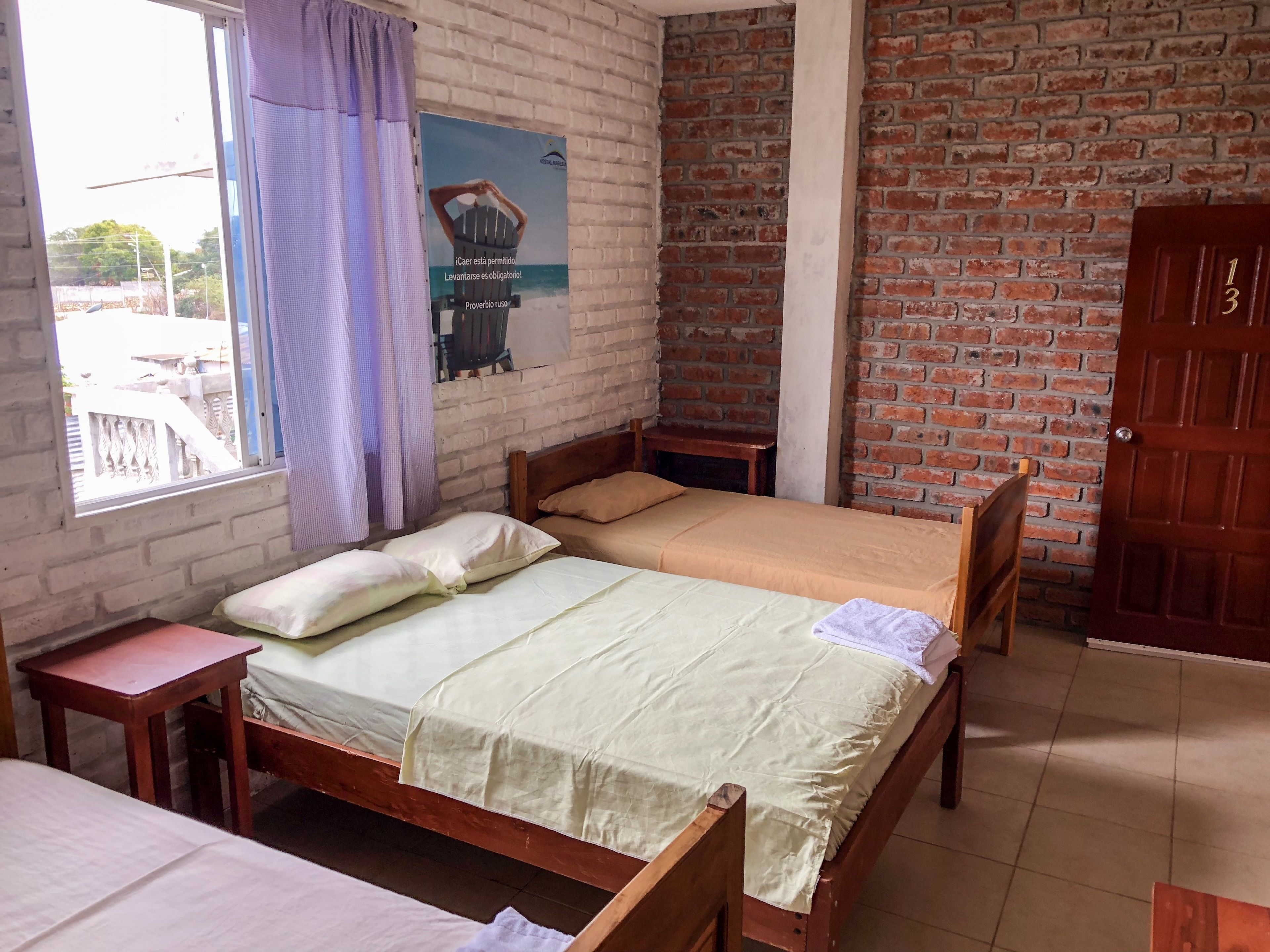Een kamer met een houten bed, een klein houten tafeltje, een raam met gordijnen, een bakstenen muur en een deur.