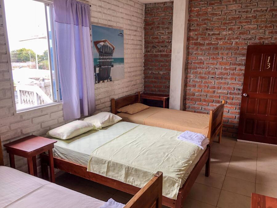 Een kamer met een houten bed, een klein houten tafeltje, een raam met gordijnen, een bakstenen muur en een deur.