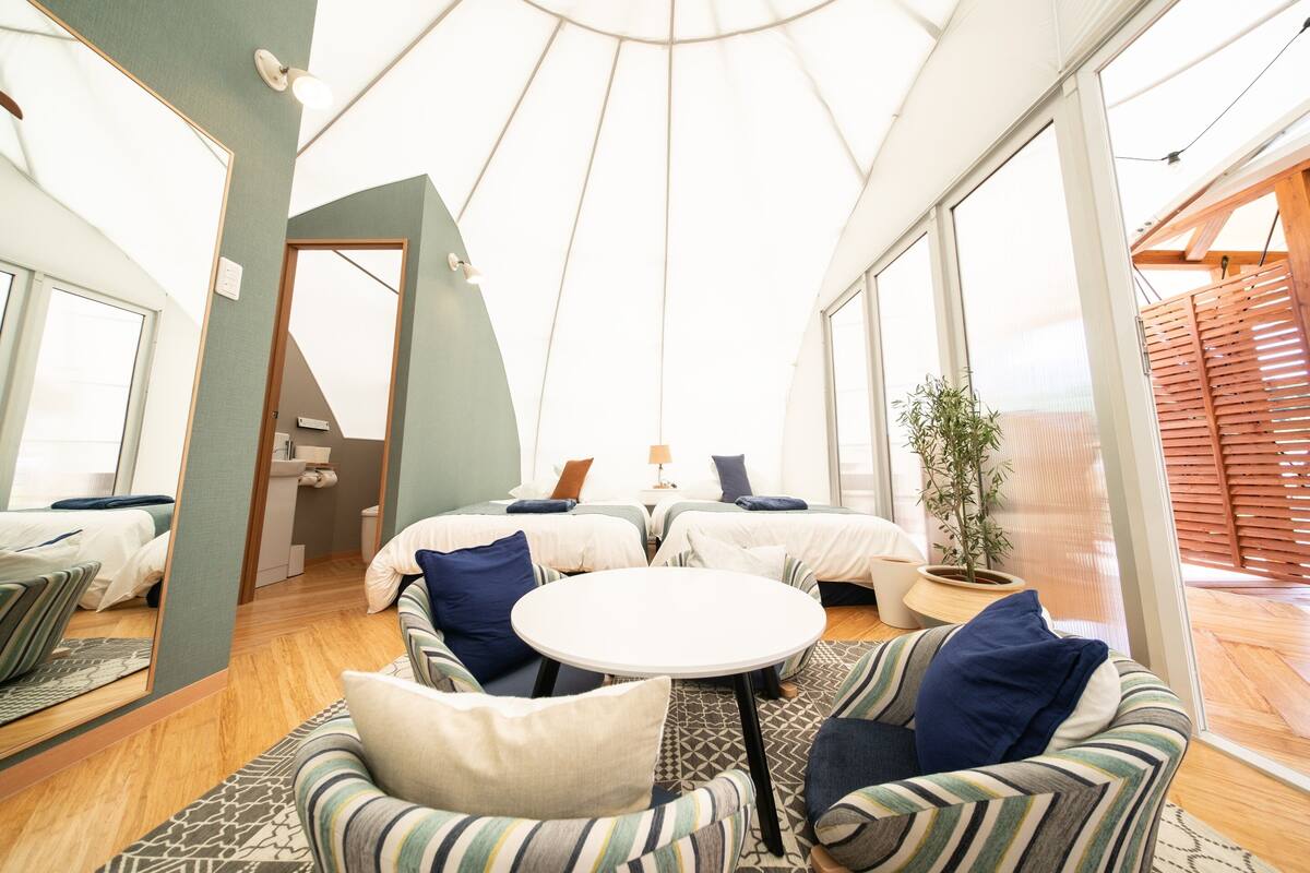 A modern, spacious room with a high ceiling and large windows, featuring a central seating area with sofas and a round table, and a visible bathroom with a toilet and sink.
