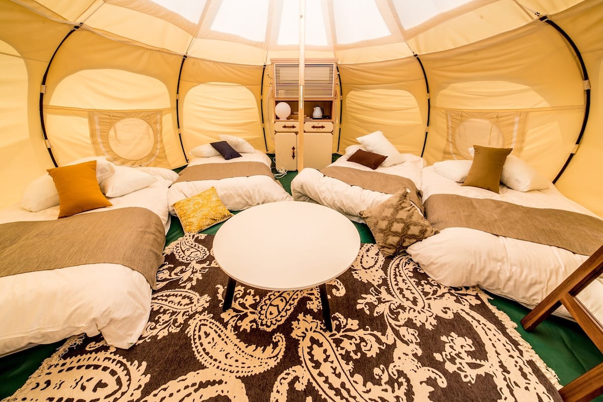 A cozy interior of a dome-shaped tent with a central round table, multiple beds with pillows, and a patterned rug.