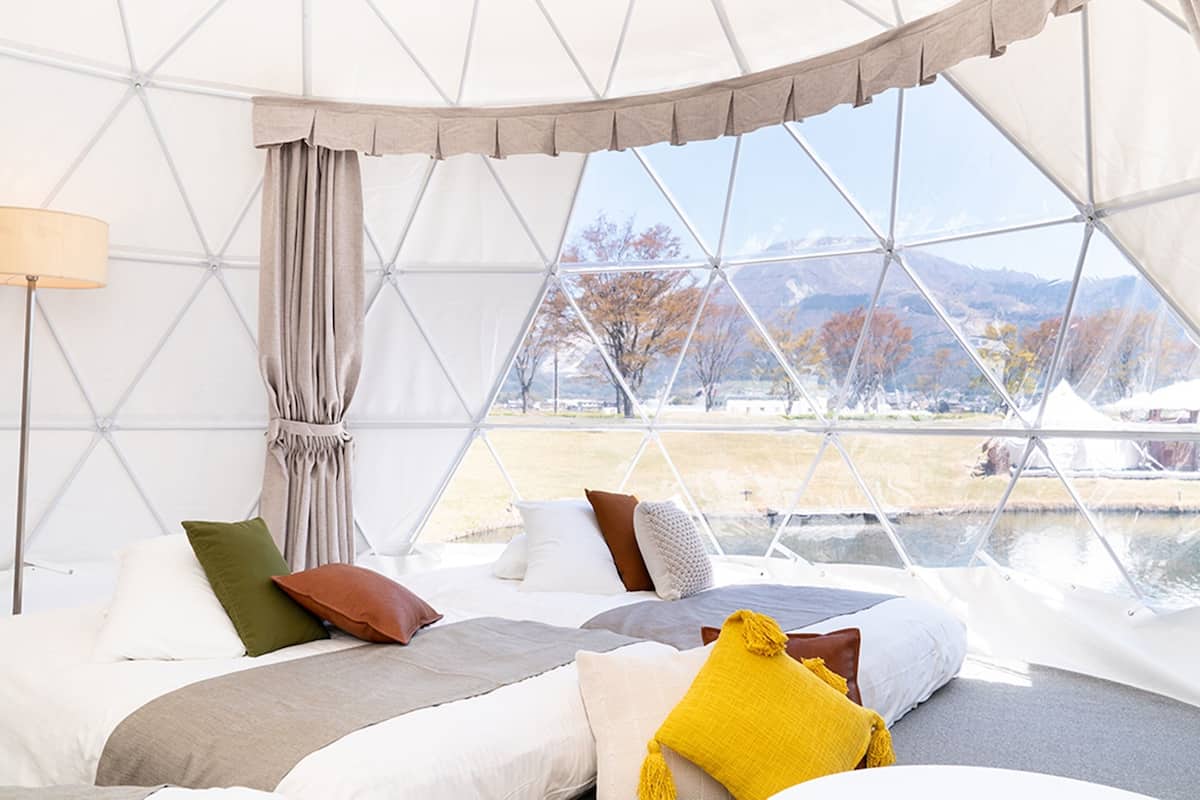A spacious room with a bed, pillows, a lamp, and a view of a landscape through large windows.