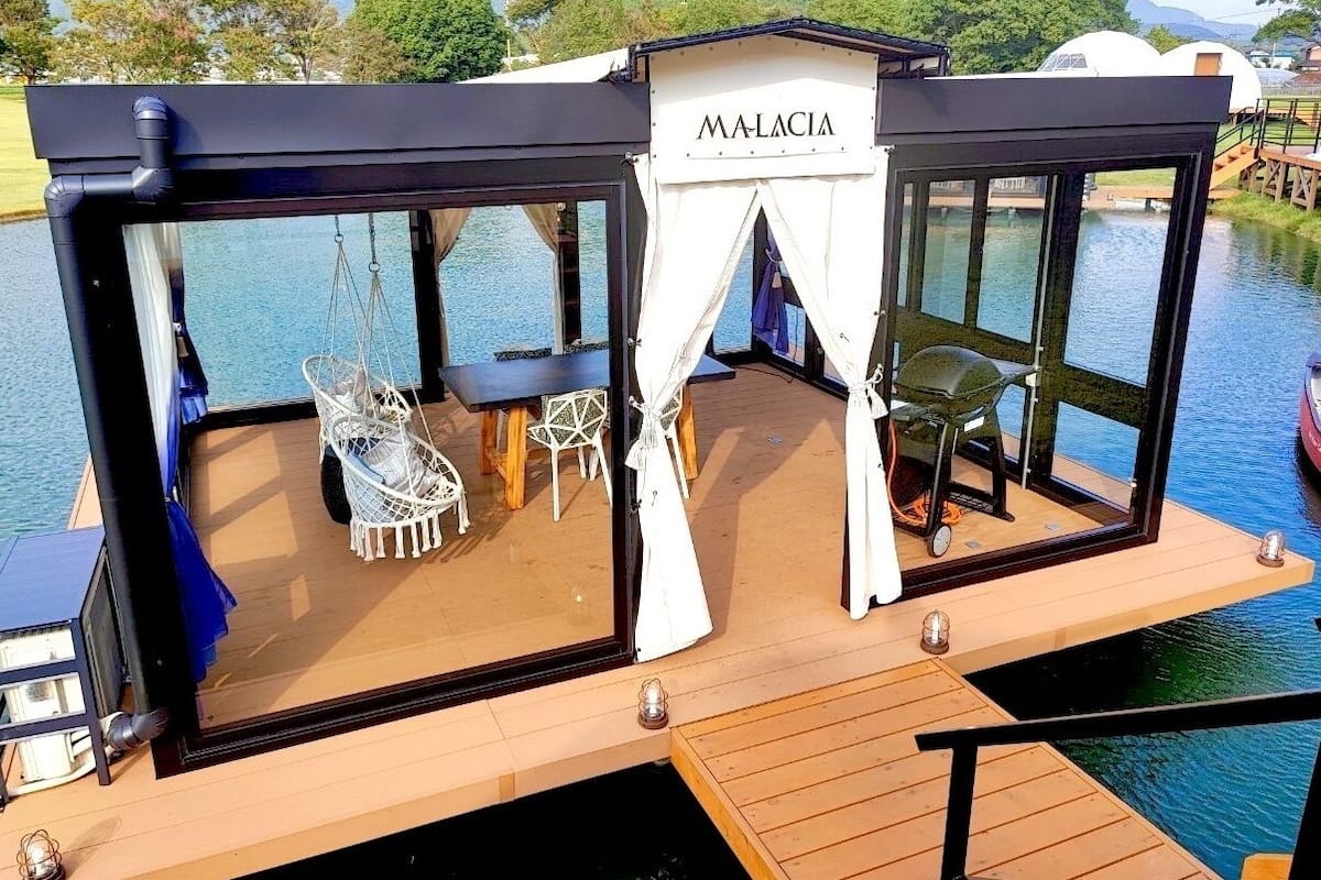 A floating structure with a name sign 'MALACIA', featuring a dining area with white furniture and a view of the water.