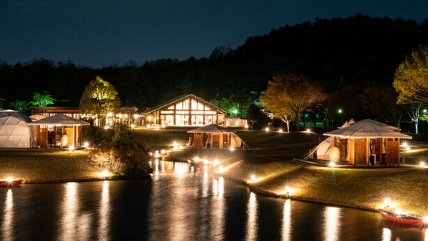 Un resort junto al lago con varias tiendas de campaña y un albergue de madera, rodeado de árboles e iluminado por luces nocturnas.