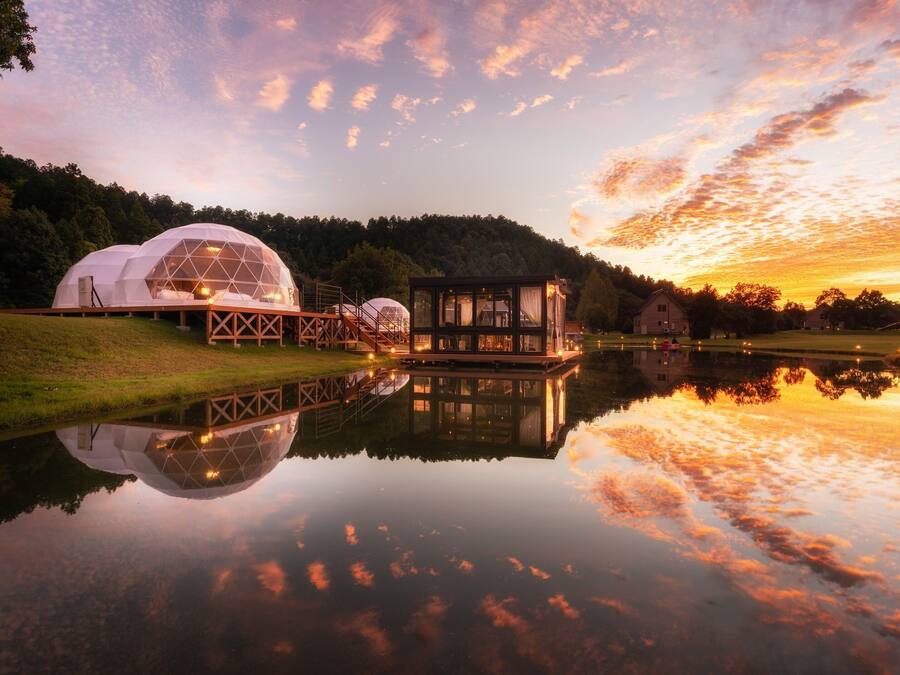 A lakeside view of geodesic dome accommodations with a reflection in the water, surrounded by trees and a sunset sky.
