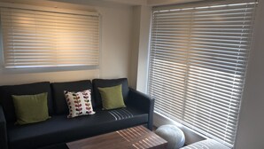 A modern living room with a dark-colored sofa, two green cushions, a wooden coffee table, and a large window with blinds. - Condominium MIRAHAKONE (Hakone)