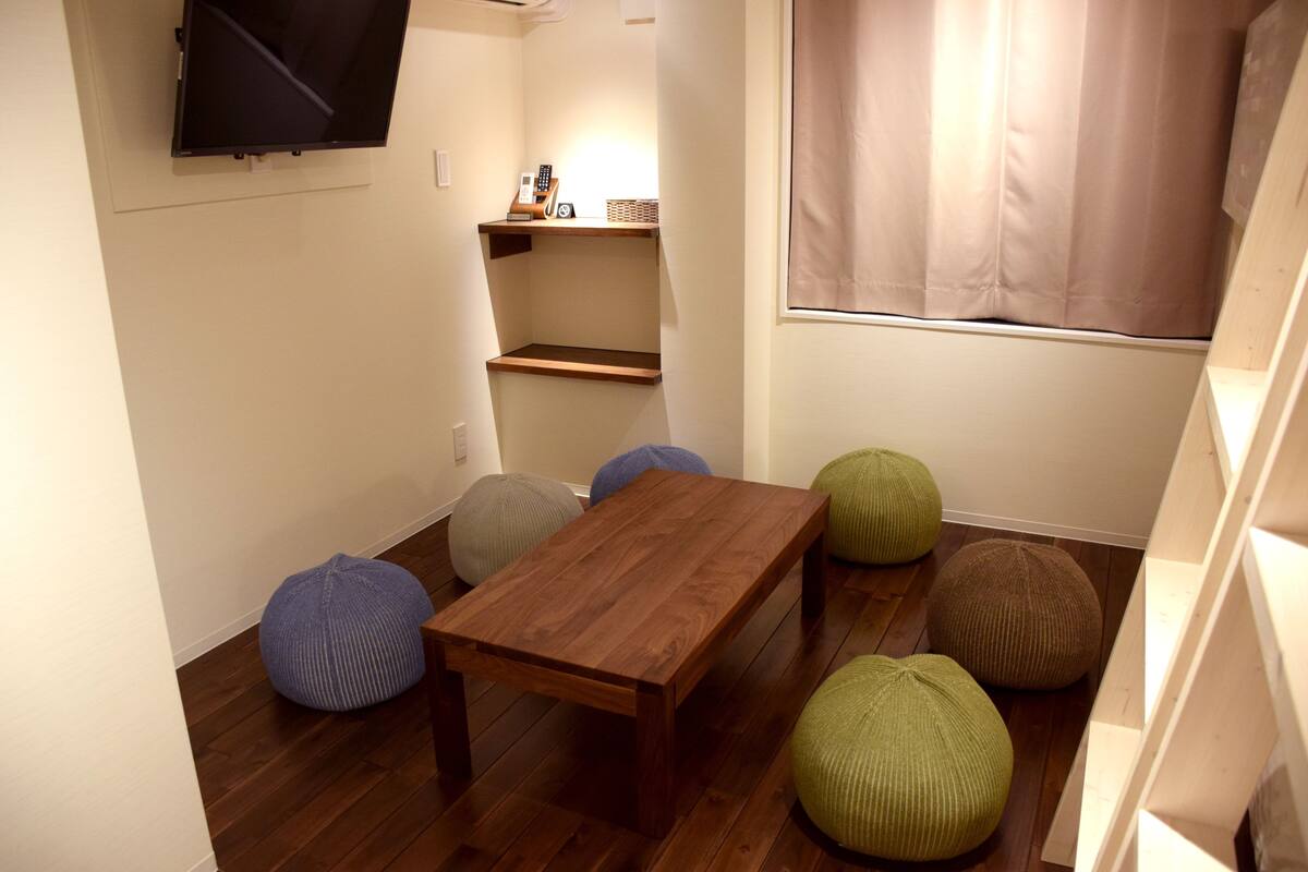 Ein kleiner Raum mit einem Holztisch, vier Sitzsäcken, einem an der Wand montierten Fernseher und einem Fenster mit Vorhang.