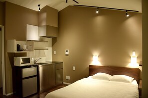A compact hotel room with a kitchenette, a bed with white linens, and two wall-mounted reading lights.