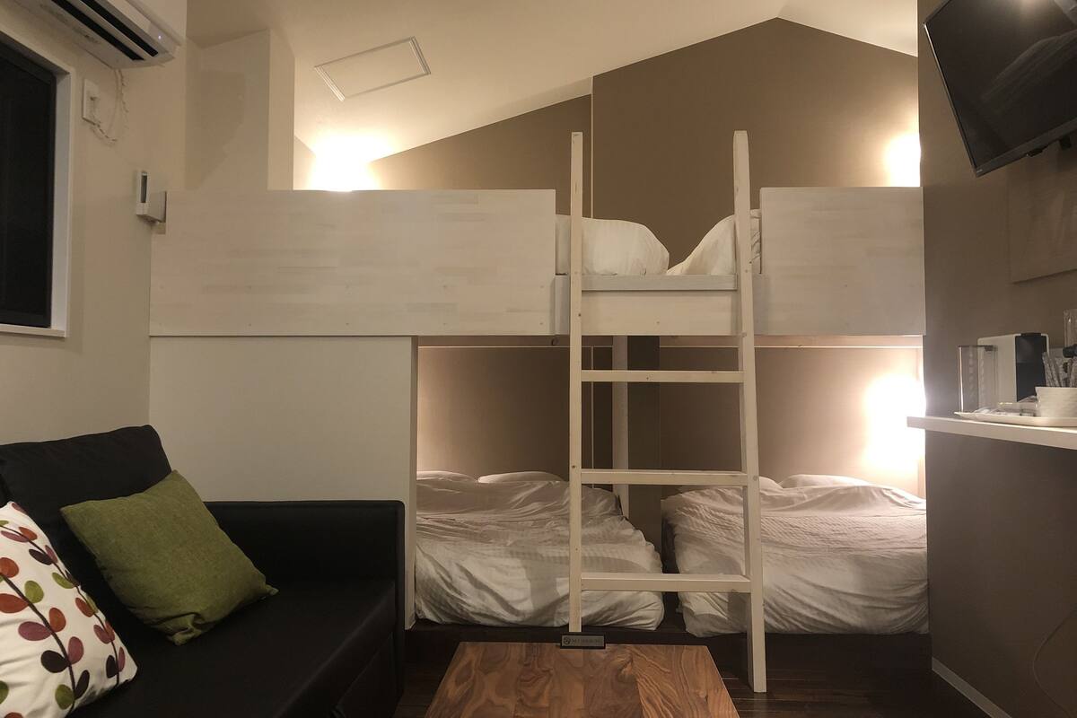 Ein Zimmer mit Etagenbetten, einer schwarzen Schlafsofa mit gepunktetem Kissen, einem Holztisch und einem an der Wand befestigten Fernseher.