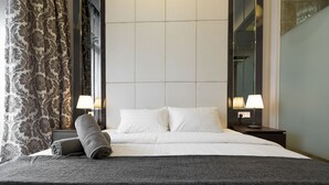 Een net opgemaakt bed met witte lakens en een grijze deken, twee bedlampen en een kamerbreed gordijn.