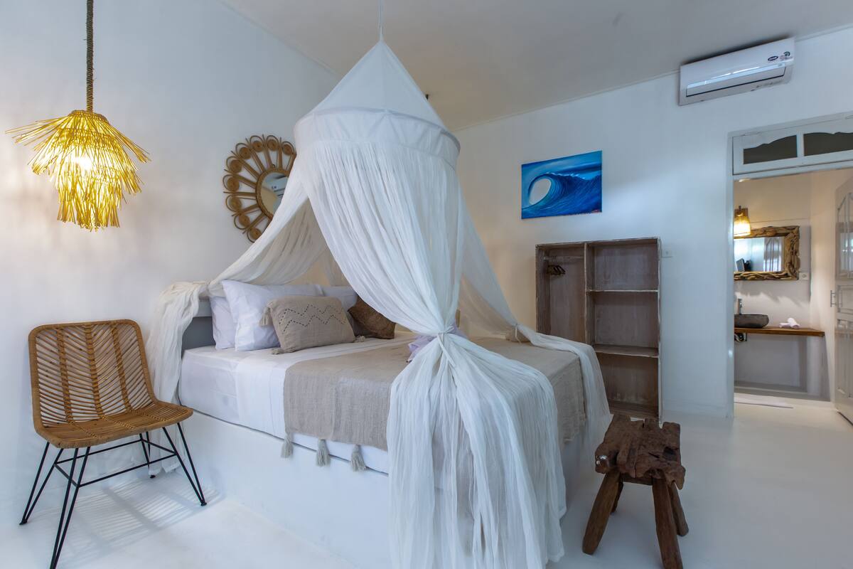 Een slaapkamer met een hemelbed, een rieten stoel, een houten kruk en een spiegel aan de muur.