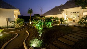 Un chemin bien éclairé mène à un bâtiment dont le toit est recouvert de tuiles, entouré de jardins aménagés et de palmiers.