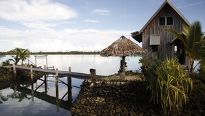 A wooden house with a thatched roof by a calm lake, a dock extending into the water, and a stone wall. - Serah's Lagoon Hideaway (Auki)