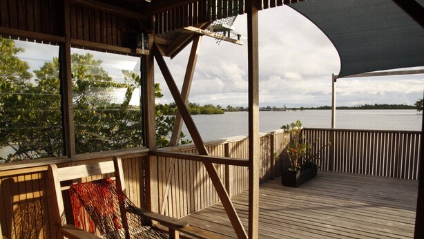 A wooden deck with a view of a body of water, a covered seating area, and a potted plant. - Serah's Lagoon Hideaway (Auki)