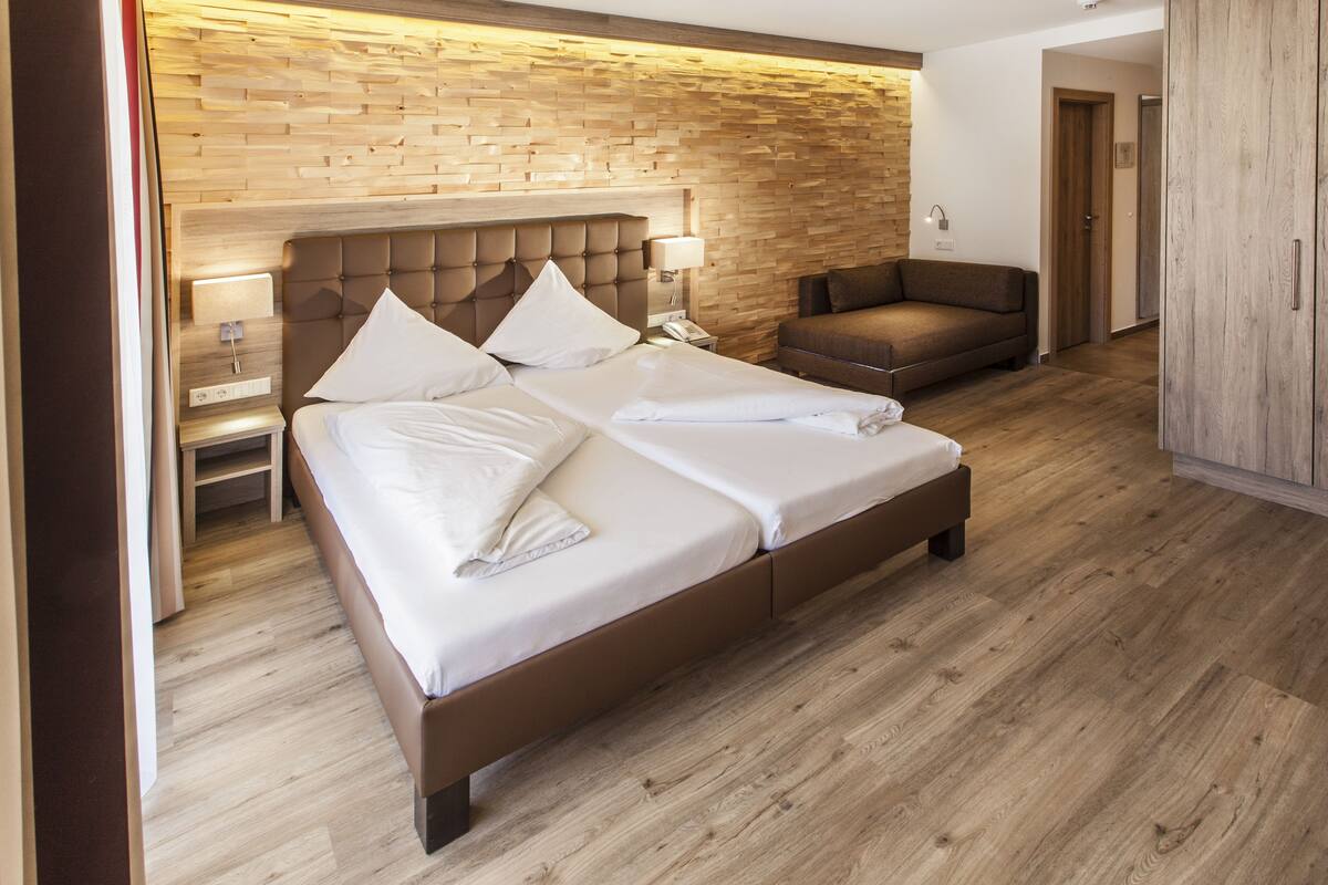 Quarto de hotel moderno com uma cama grande, um sofá, cabeceira de madeira e piso de madeira.