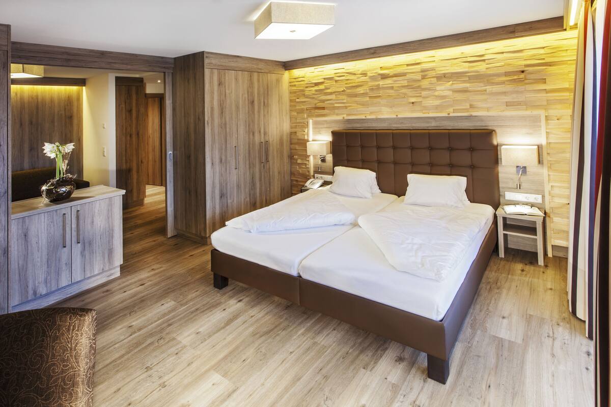Quarto de hotel moderno com uma cama grande, guarda-roupa de madeira, mesas de cabeceira e uma pequena escrivaninha com abajur.