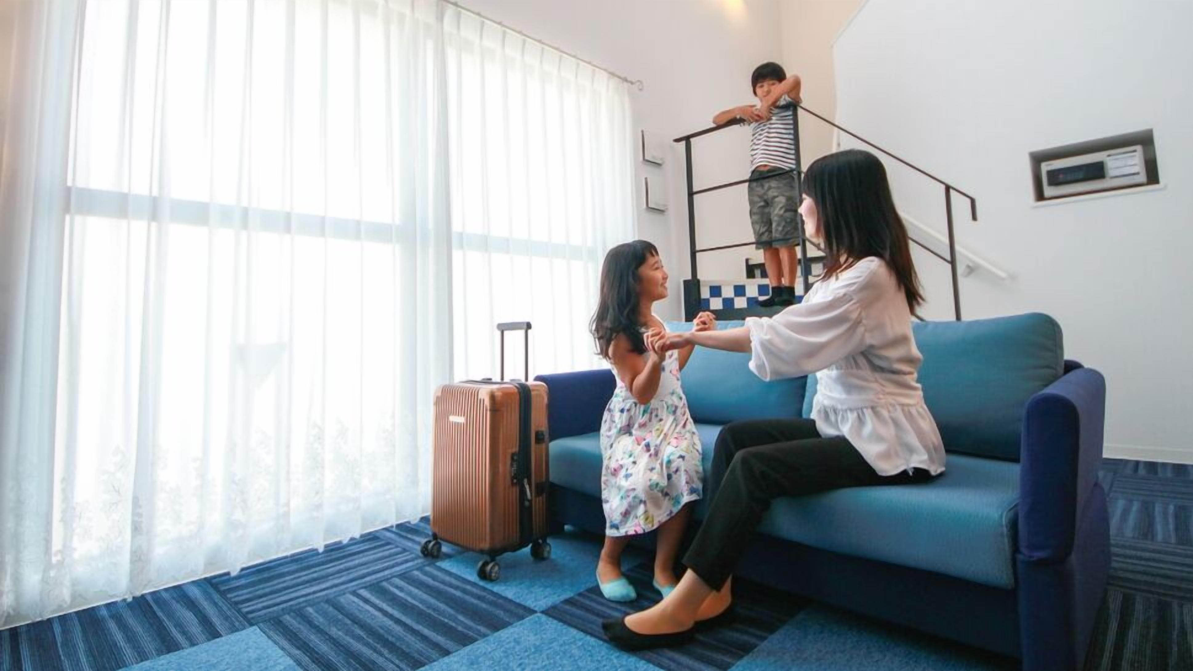 A room with a blue sofa, a child with a suitcase, and a person standing on a small platform.