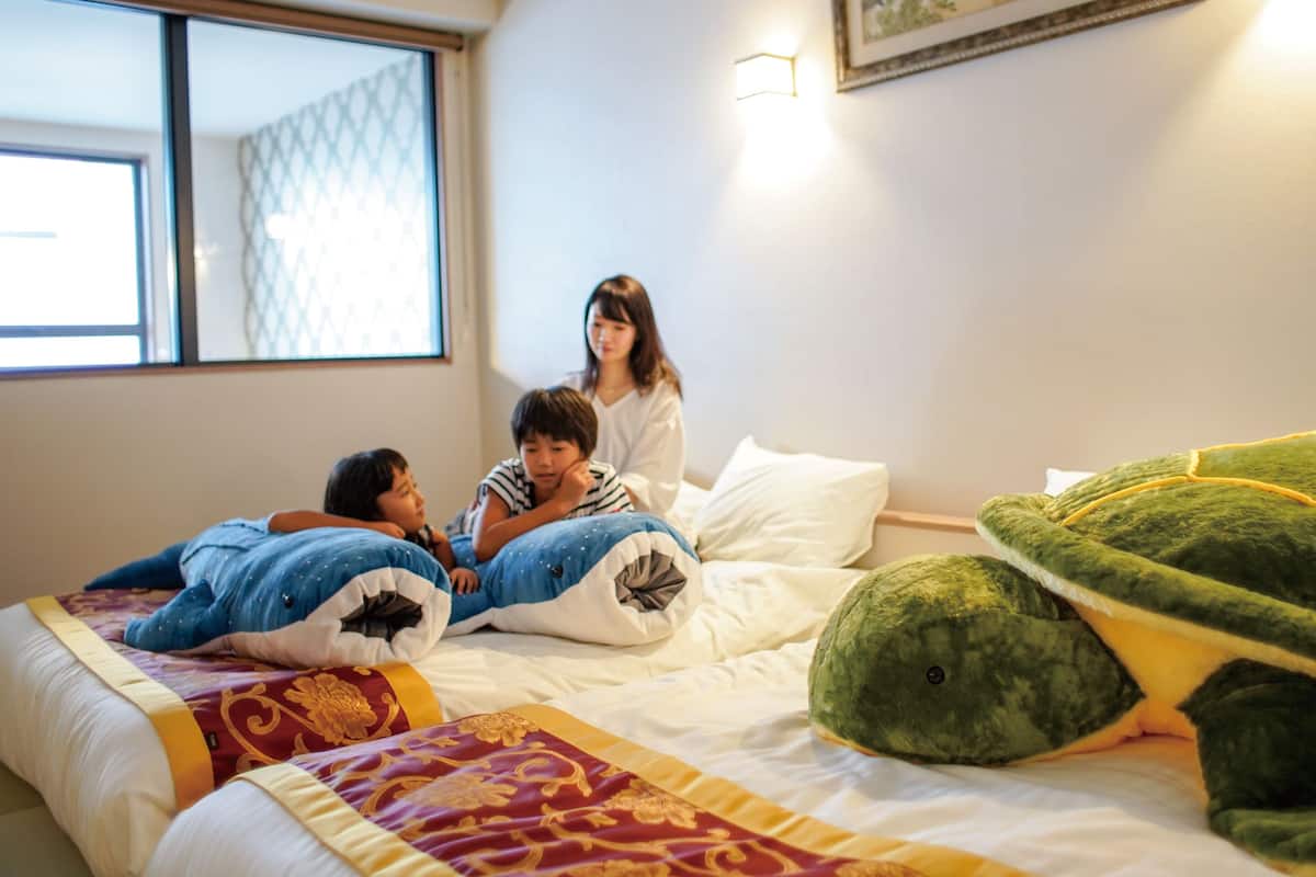 Een slaapkamer met een bed, kussens, een deken, een groen knuffeldier en een schilderij aan de muur.