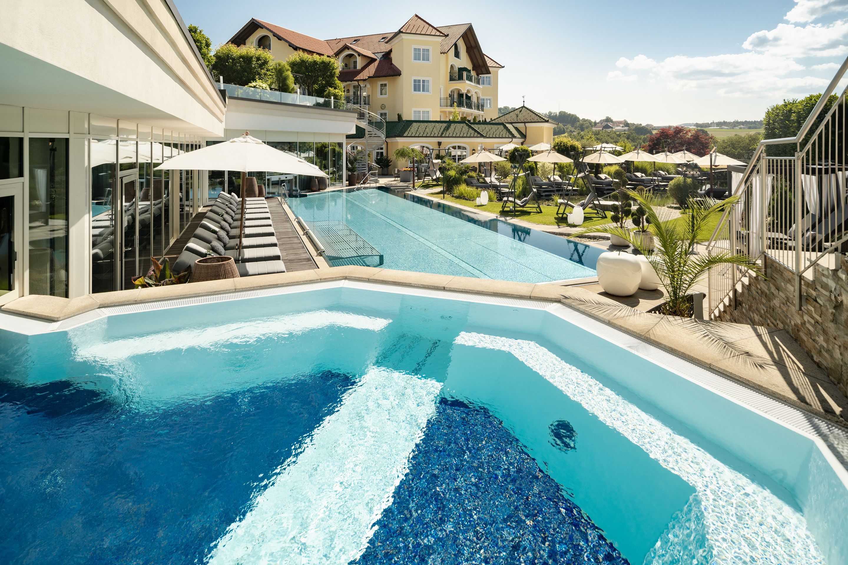 Piscina con sillones de descanso y sombrillas, rodeada de edificios y vegetación.