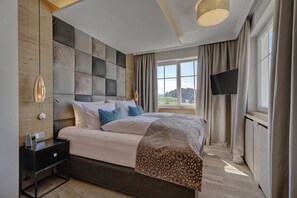 A modern bedroom with a large bed, a bedside table with a lamp, a flat-screen TV mounted on the wall, and a window with curtains.
