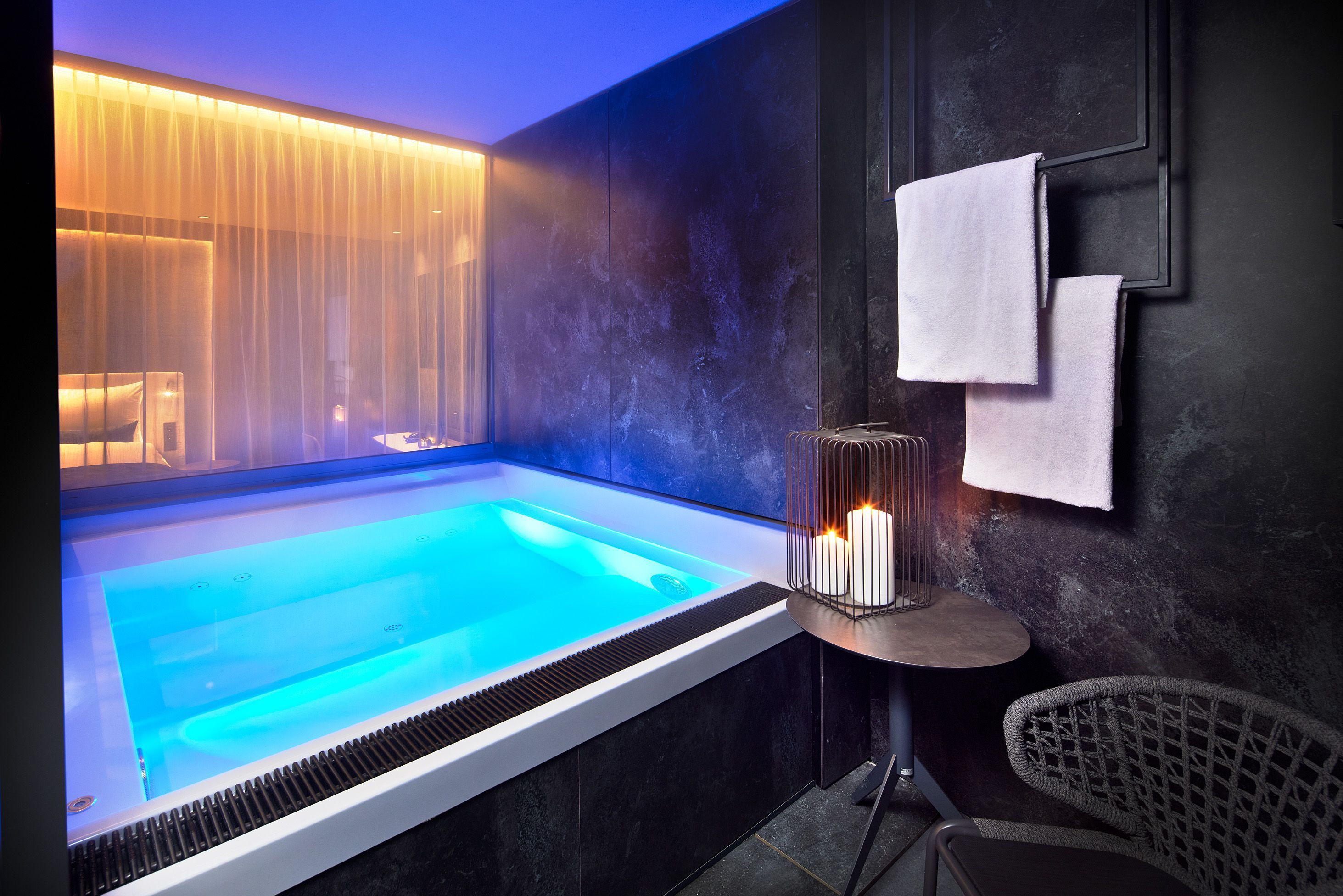 Un moderno spa con piscina climatizada, una pequeña mesa con velas y una silla de mimbre.