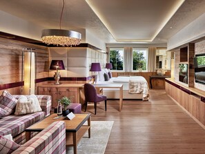 A modern hotel room with a wooden floor, a sofa, a bed, a desk, and a large window. - ROMANTIK-SUITE - 5* Wellness- & Sporthotel Jagdhof (Röhrnbach)