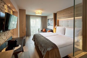 A hotel room with a large bed, a desk with a coffee machine, a TV mounted on the wall, and a window with curtains. - DOUBLE ROOM BERGKRISTALL - 5* Wellness & Sporthotel Jagdhof (Röhrnbach)